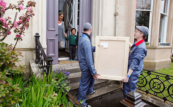 Street with mother and child standing at open front door, with two men delivering art work to them 
